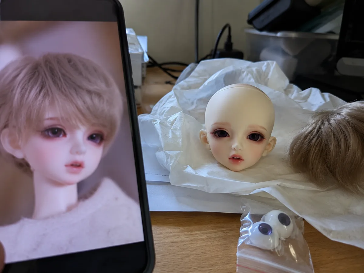 QC compare at home: A buyer compares a blurred QC reference on phone with the received head, wig, and eyes at home.